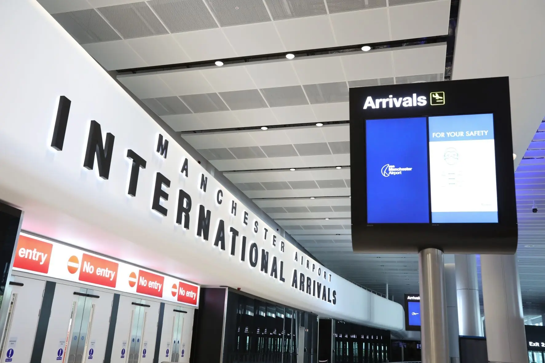 Arrivals hall at Manchester Airport with travelers collecting luggage while a chauffeur greets a passenger