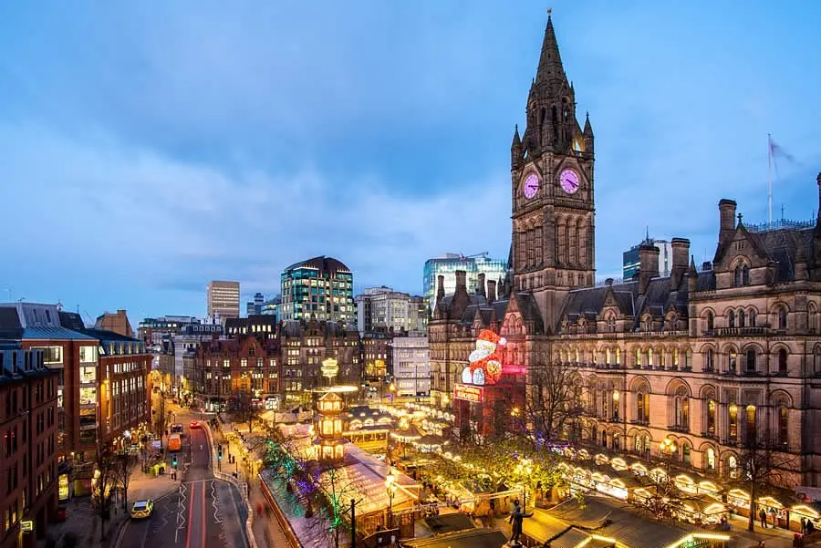 Manchester city centre at dusk featuring the historic Town Hall, illuminated streets, and festive market stalls creating a vibrant urban atmosphere